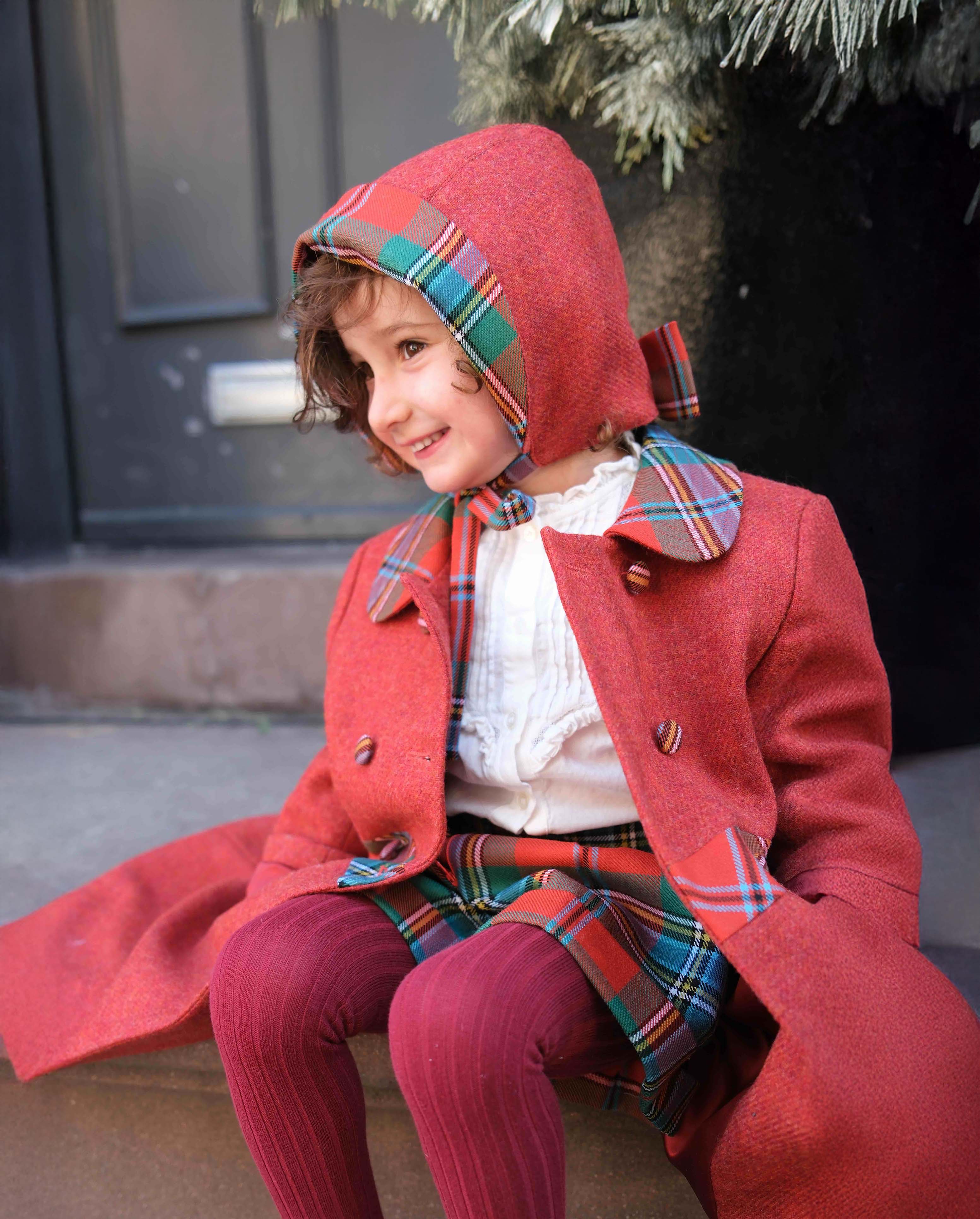 Child wearing a red coat and tartan hat sitting on a step.
